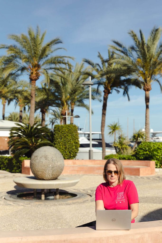 Clair of Chief Stew Shop wearing pink tee shirt and working on her laptop, with palmtrees and yachts in the background during her personal branding shoot. 